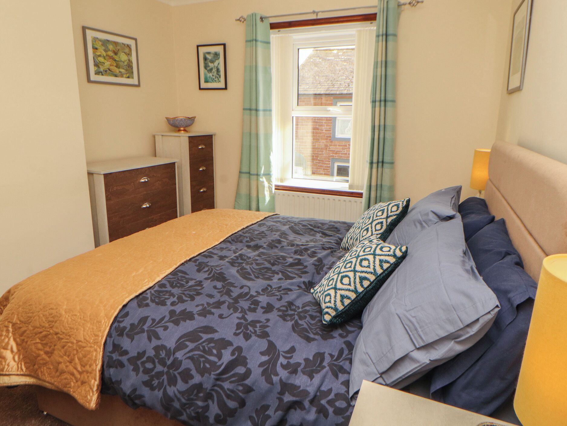 A bedroom with a bed, a dresser, a window with curtains, and two framed pictures on the wall.