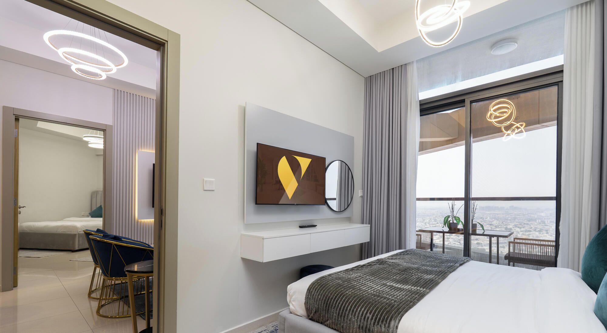 A modern hotel room with a bed, a TV mounted on the wall, a balcony with a view, and a dining area with a table and chairs.