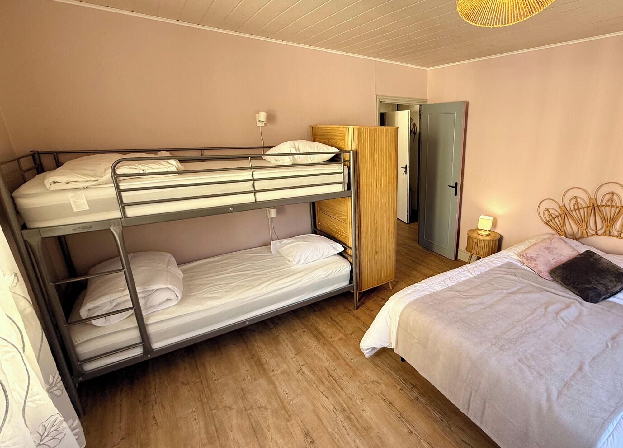 A bunk bed room with a wooden wardrobe, a bedside table with a lamp, and a door leading to another room.