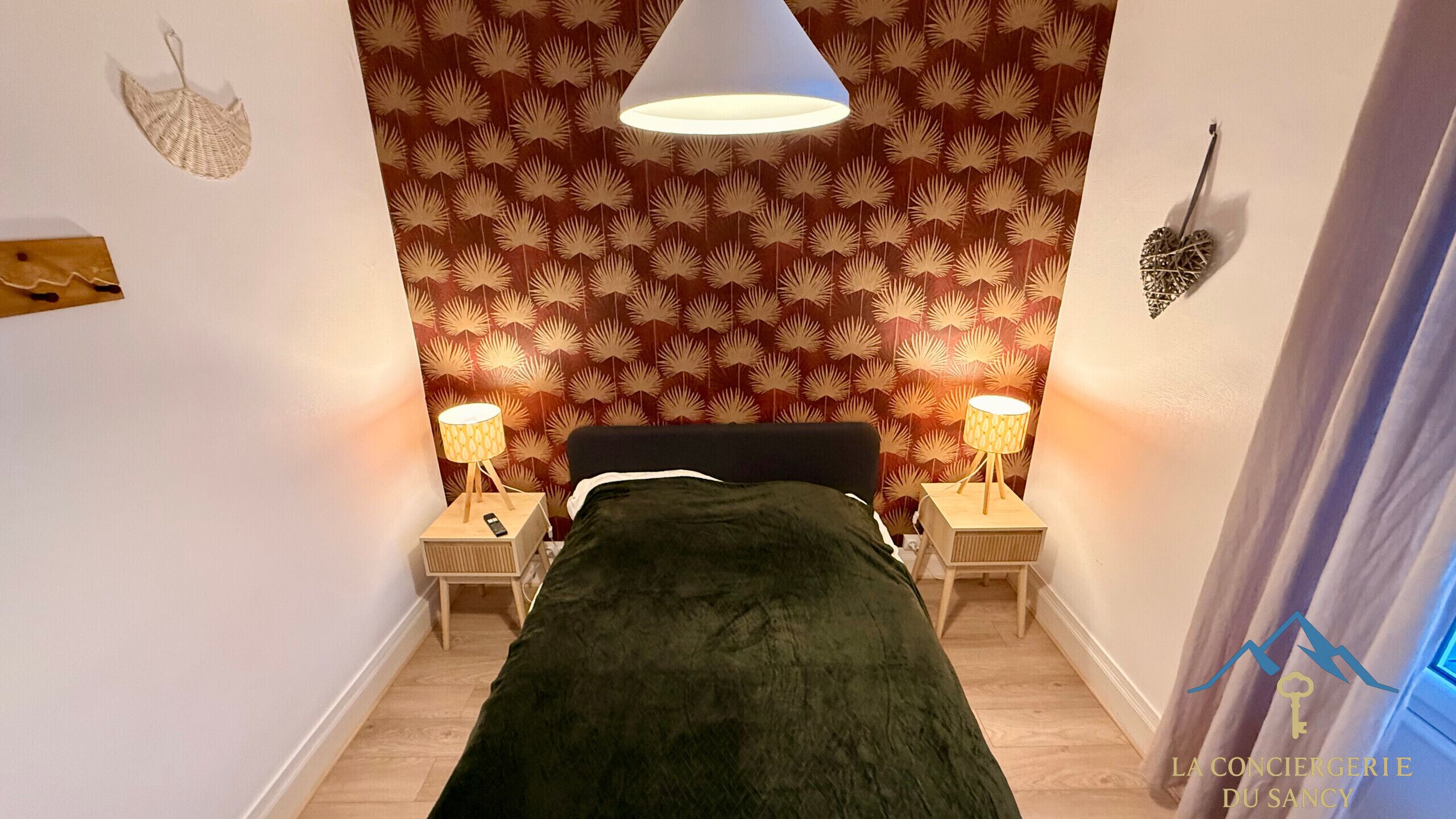 A bedroom with a bed, two bedside tables with lamps, a patterned wallpaper, and a hanging light fixture.