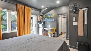 A bedroom with a bed, a desk with chairs, a TV, and a fan.