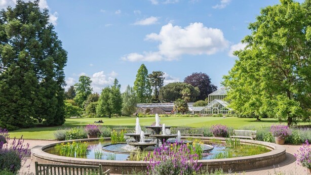 Ein Garten mit Brunnen, Bänken und einem Gewächshaus im Hintergrund.