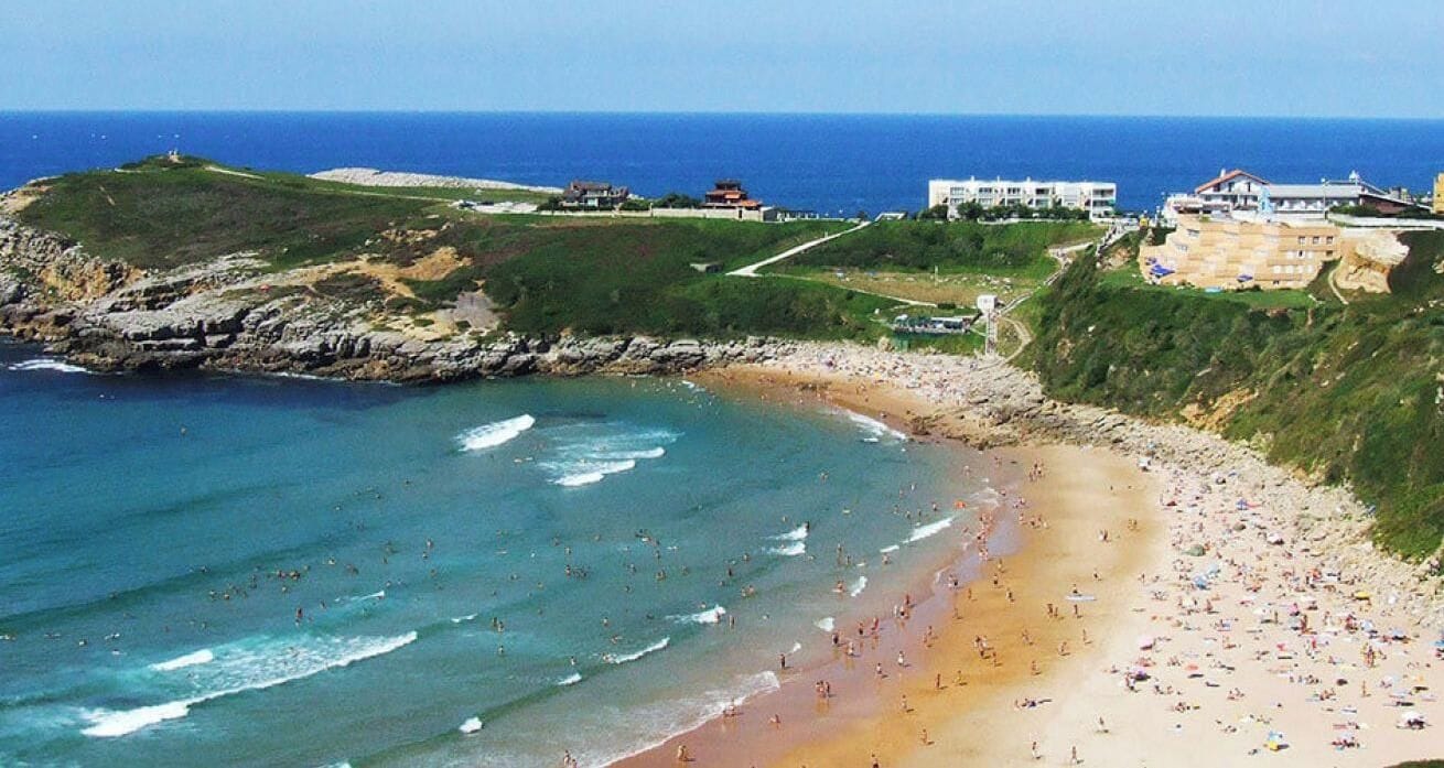 Une plage avec un littoral rocheux, une étendue de sable et des bâtiments en arrière-plan.