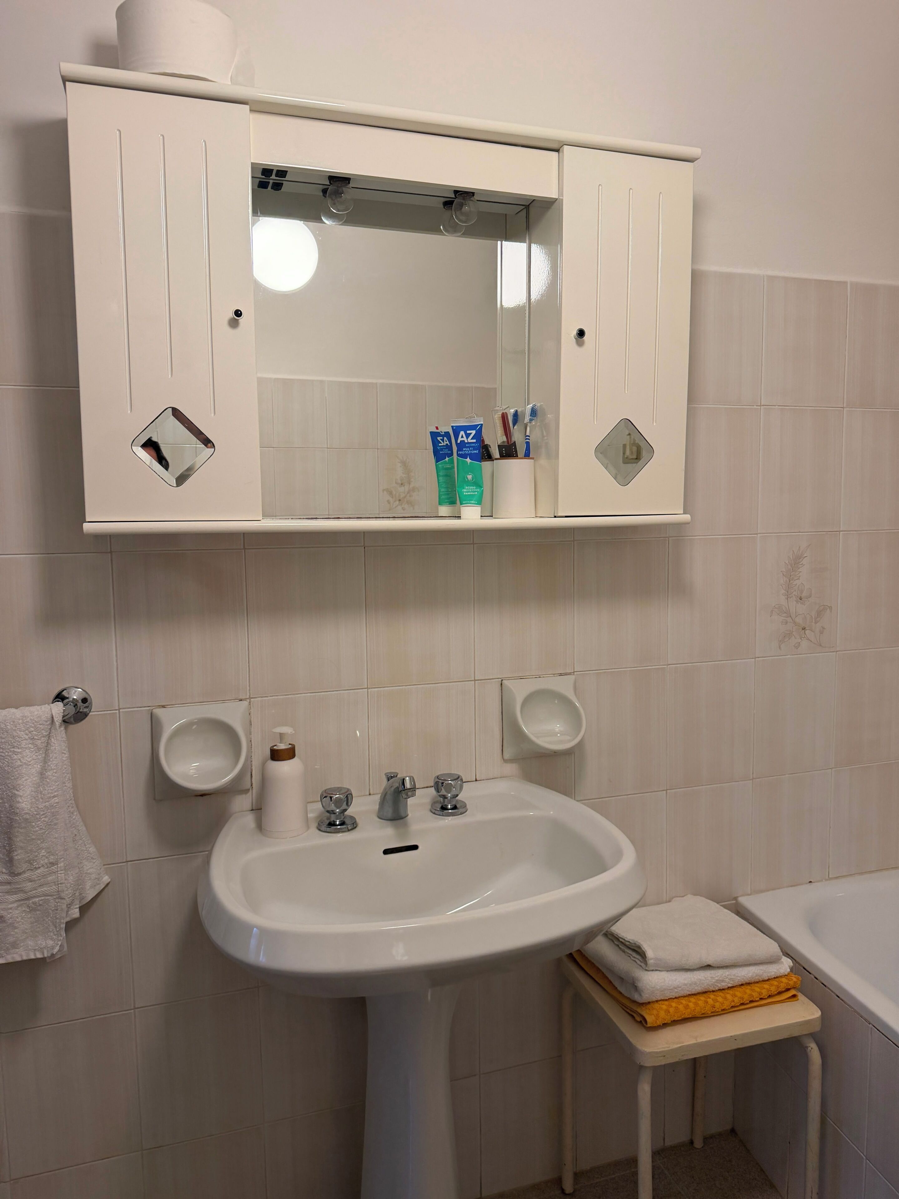 Un baño con lavamanos blanco, espejo y mueble, un perchero y una mesita con toallas.