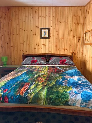 A wooden-paneled room with a bed featuring a colorful landscape bedspread, a green nightstand, and a framed picture on the wall.