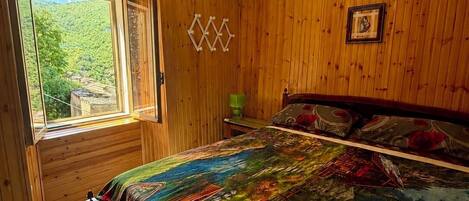 Una habitación con paredes de madera, una cama con colcha colorida, una ventana con vistas al verdor y una lámpara en el techo.