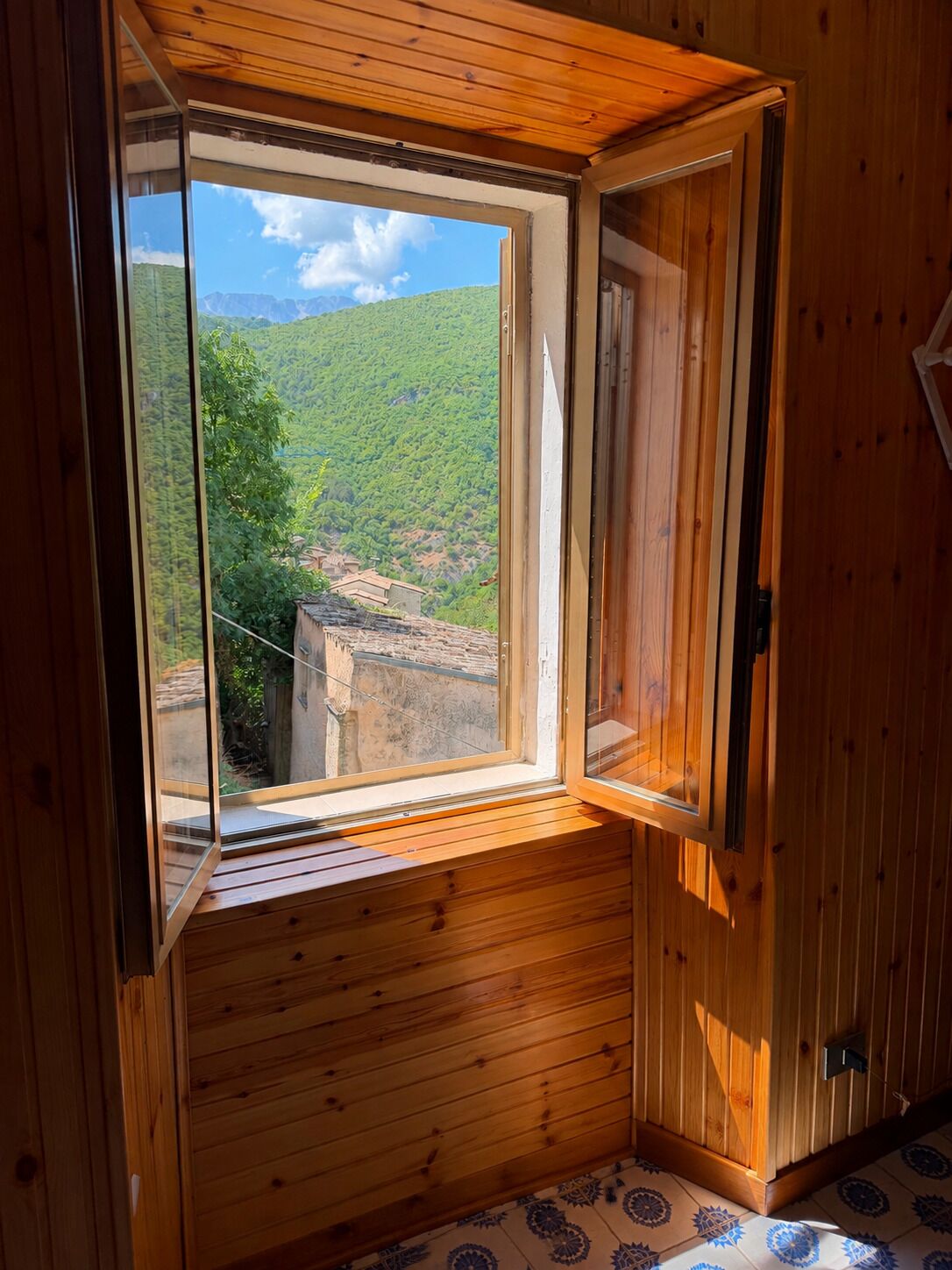 Quarto de madeira com uma janela grande que oferece vista para uma paisagem verde e edifícios ao longe.