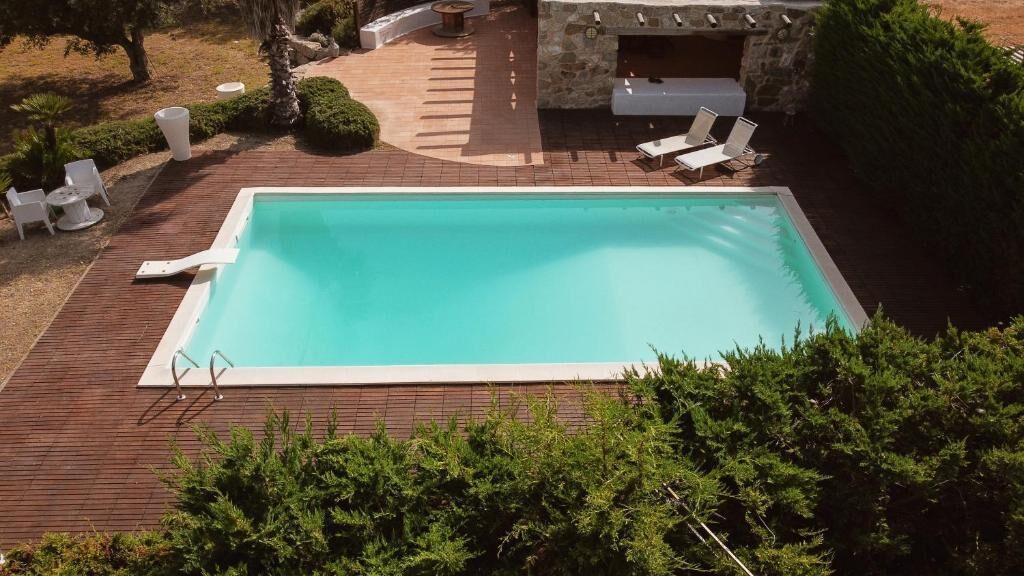 Ein rechteckiger Swimmingpool mit Holzterrasse, umgeben von Grünpflanzen und einer Steinstruktur mit Sitzgelegenheiten.