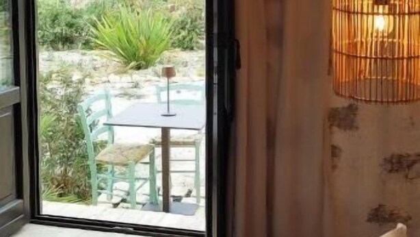 Ein Zimmer mit einem Bett, einem Vorhangfenster, einer Korblampe und Blick auf eine Terrasse mit Stühlen und Pflanzen.