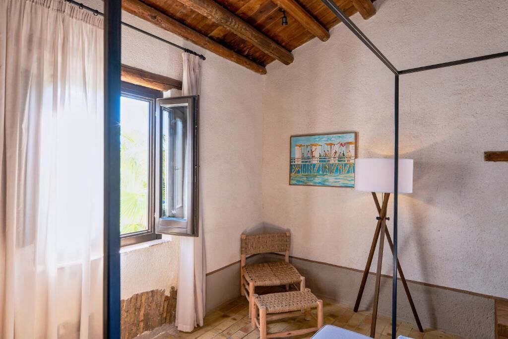 Ein Zimmer mit Holzboden, einem mit Vorhängen ausgezogenen Fenster, einem Korbstuhl, einem Bild an der Wand und einer Stehlampe.