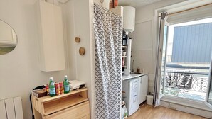 Una stanza con un comò in legno, un termosifone, un tavolino con delle bottiglie, una tenda da doccia decorata e una finestra con vista su un balcone.
