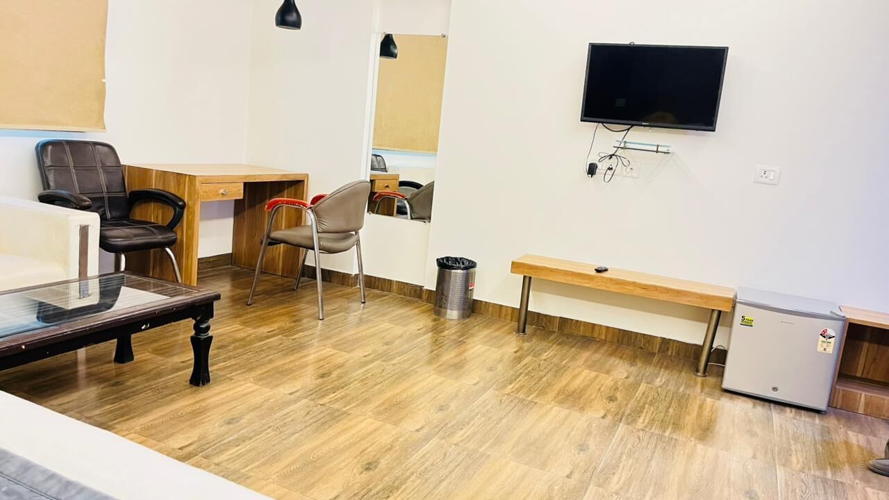 A room with a wooden desk, two chairs, a small refrigerator, a trash bin, and a mounted television.