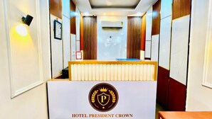 A hotel reception area with a reception desk, a logo, and a wall-mounted air conditioning unit. - Hotel President Crown (Greater Noida)
