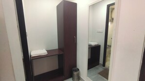 A small, enclosed bathroom with a dark wooden cabinet, a small shelf, a trash bin, and a mirror on the wall. - Raya Raj Anugraha (Kumbakonam)