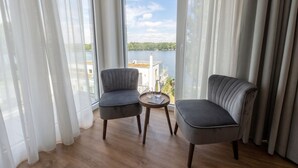A room with two grey upholstered chairs, a small round table with a glass, and a view of a lake through sheer curtains.