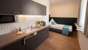 A compact hotel room with a kitchenette, a bed, and a bedside table.