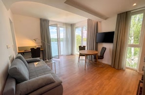 A modern living room with a sofa, a dining table, and a TV mounted on the wall. - H24 Hotel Berlin Grünau (Berlin)