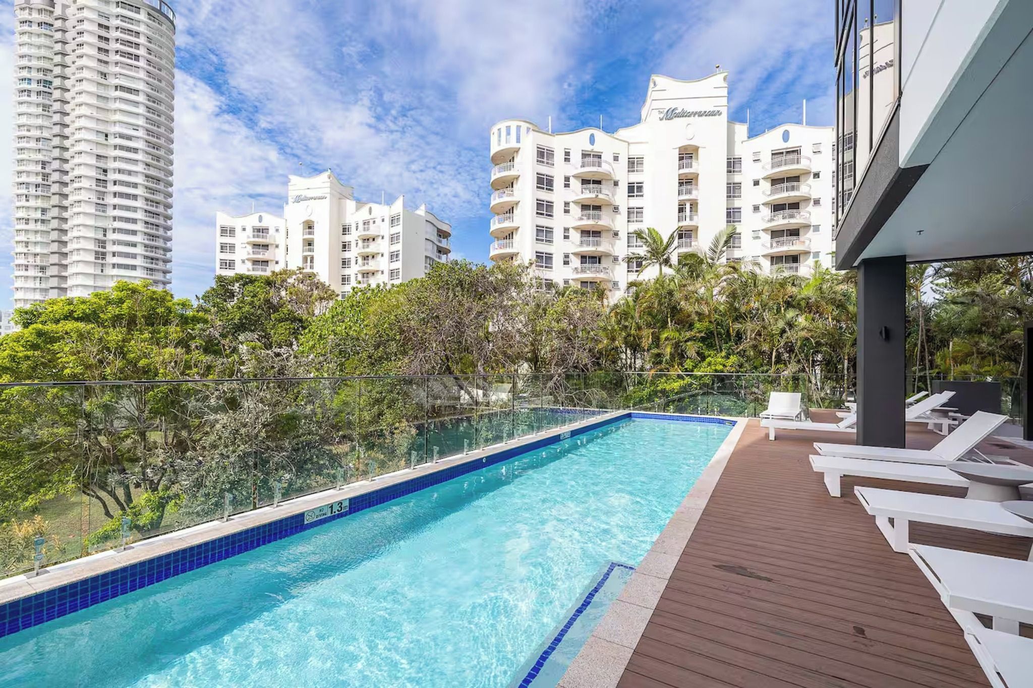 A swimming pool with a wooden deck and lounge chairs, surrounded by greenery and tall buildings.
