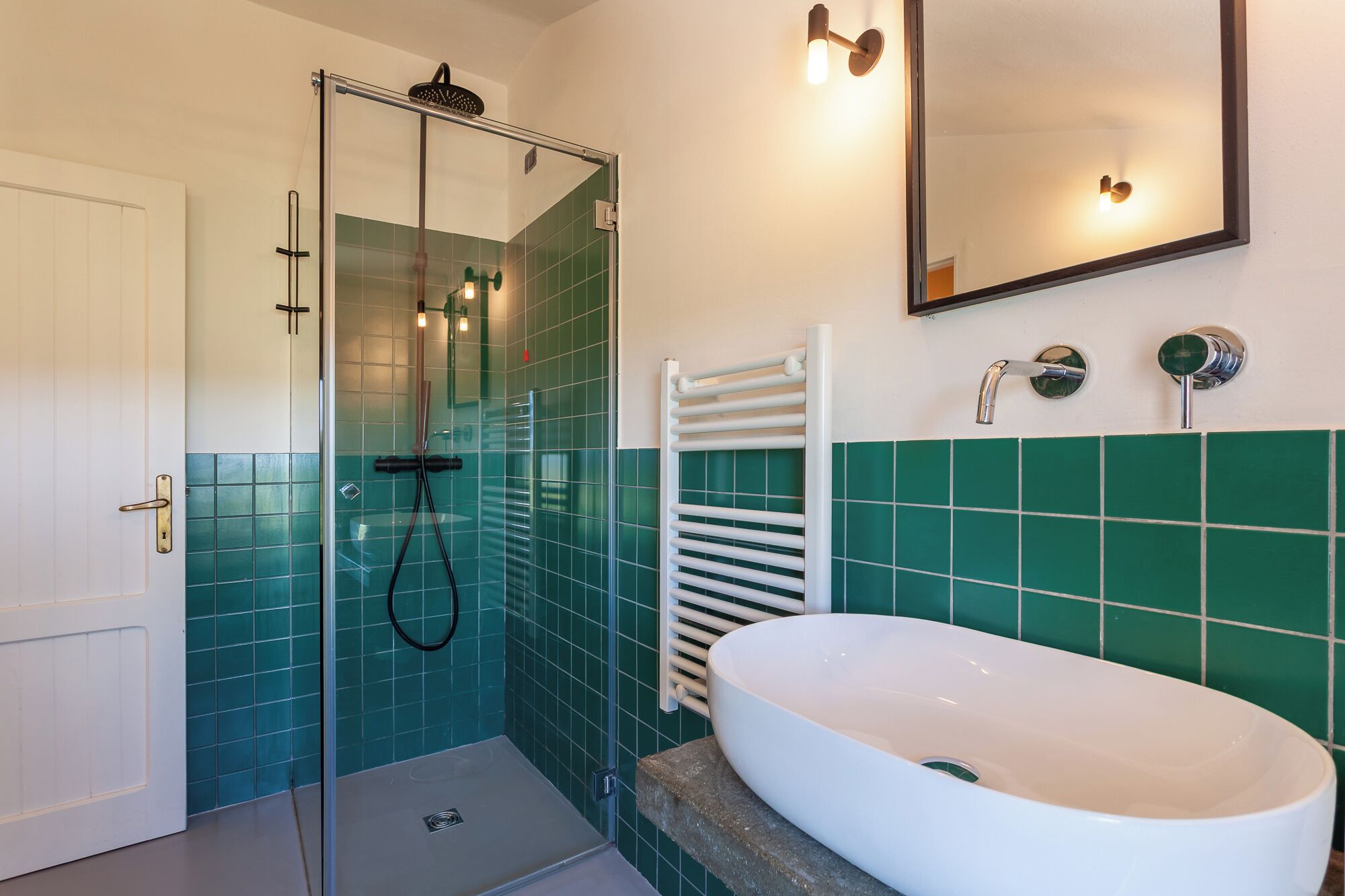 Un baño con ducha de mampara de vidrio, un lavamanos blanco y paredes cubiertas de azulejos verdes.