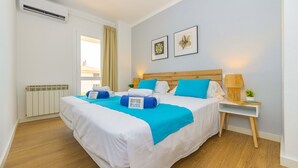 A hotel room with a large bed, a bedside table with a lamp, a window with curtains, and a radiator. - Seafront Cala Ratjada Apartment (Cala Ratjada)