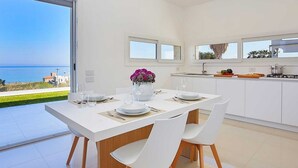 Une cuisine moderne avec des meubles blancs, un coin repas doté d’une table et de chaises blanches, et une vue sur la mer.