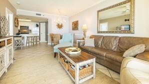 Ein Wohnzimmer mit braunem Ledersofa, einem Holztisch, Korbwannen, einem blauen Ohrensessel, einem weißen Sofa und einem großen Spiegel mit goldener Rahmung. Im Hintergrund ist eine Küche zu sehen.
