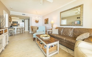 A living room with a brown leather sofa, a wooden coffee table, wicker baskets, a blue patterned armchair, a white couch, a large mirror with a gold frame, and a kitchen in the background. - Phoenix VIII by Brett Robins Vacations (Orange Beach)