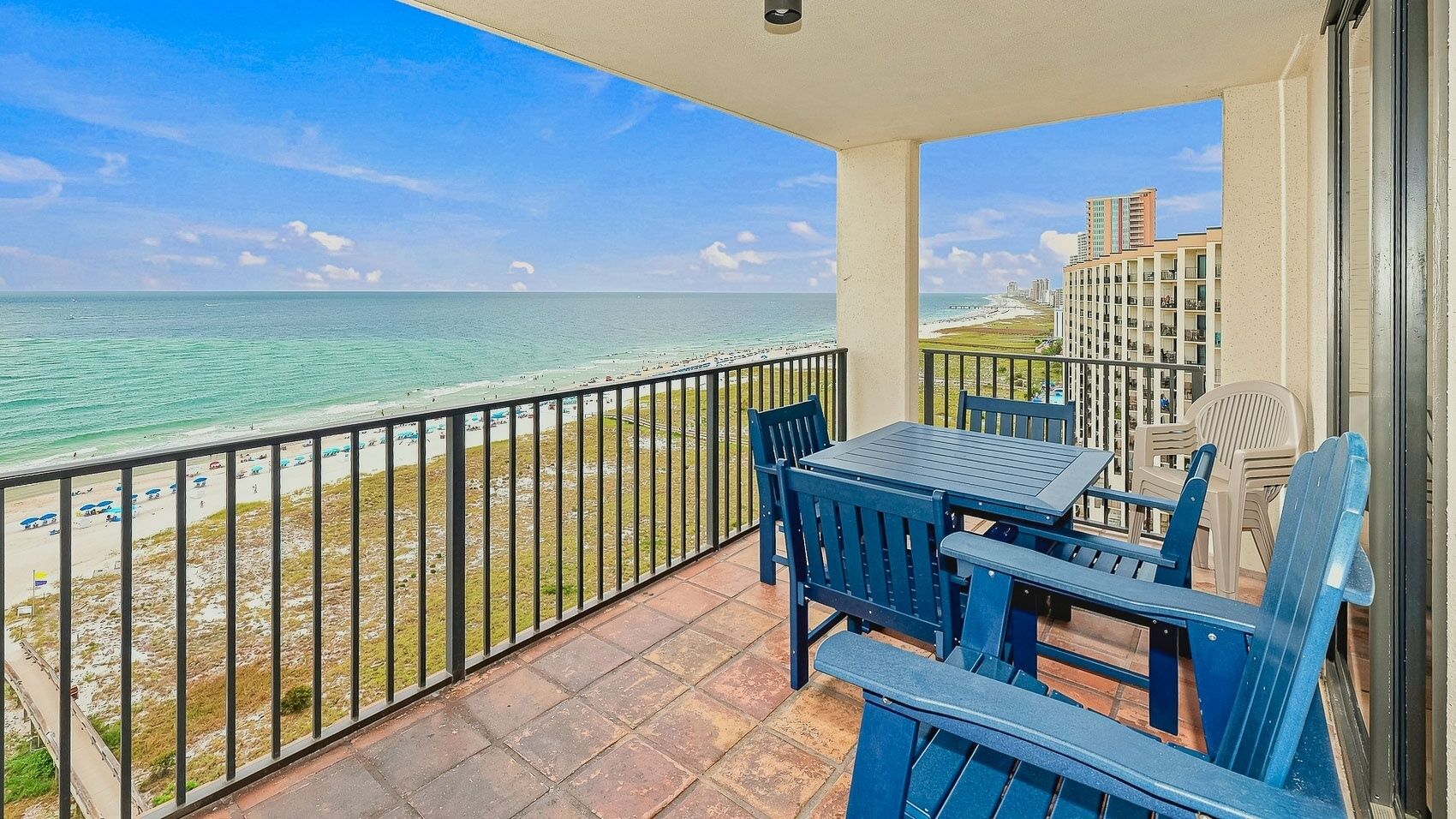 A balcony with a view of the ocean and beach, a table with chairs, and a building in the background.