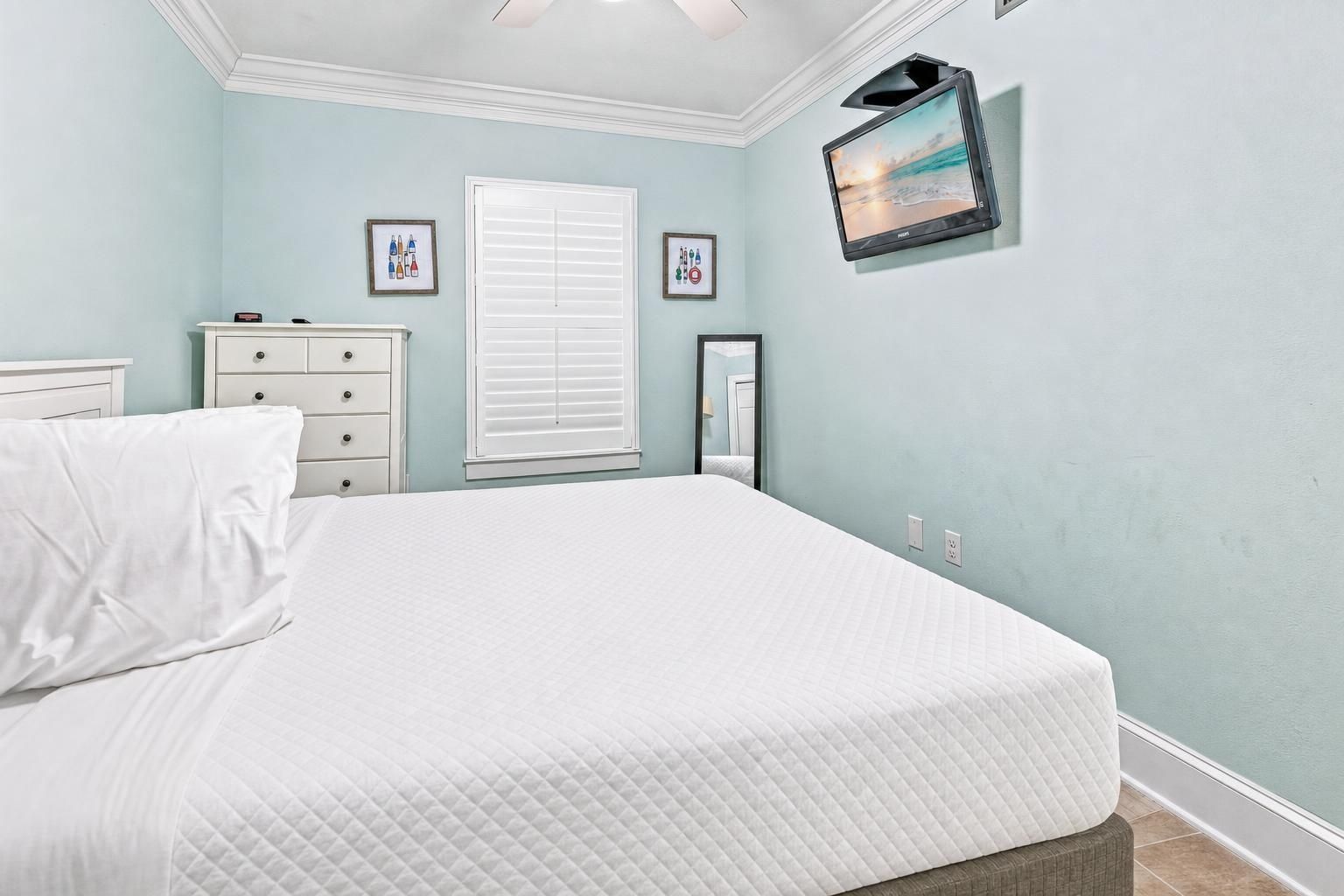 A bedroom with a bed, a dresser, a window with blinds, a wall-mounted TV, and framed pictures.