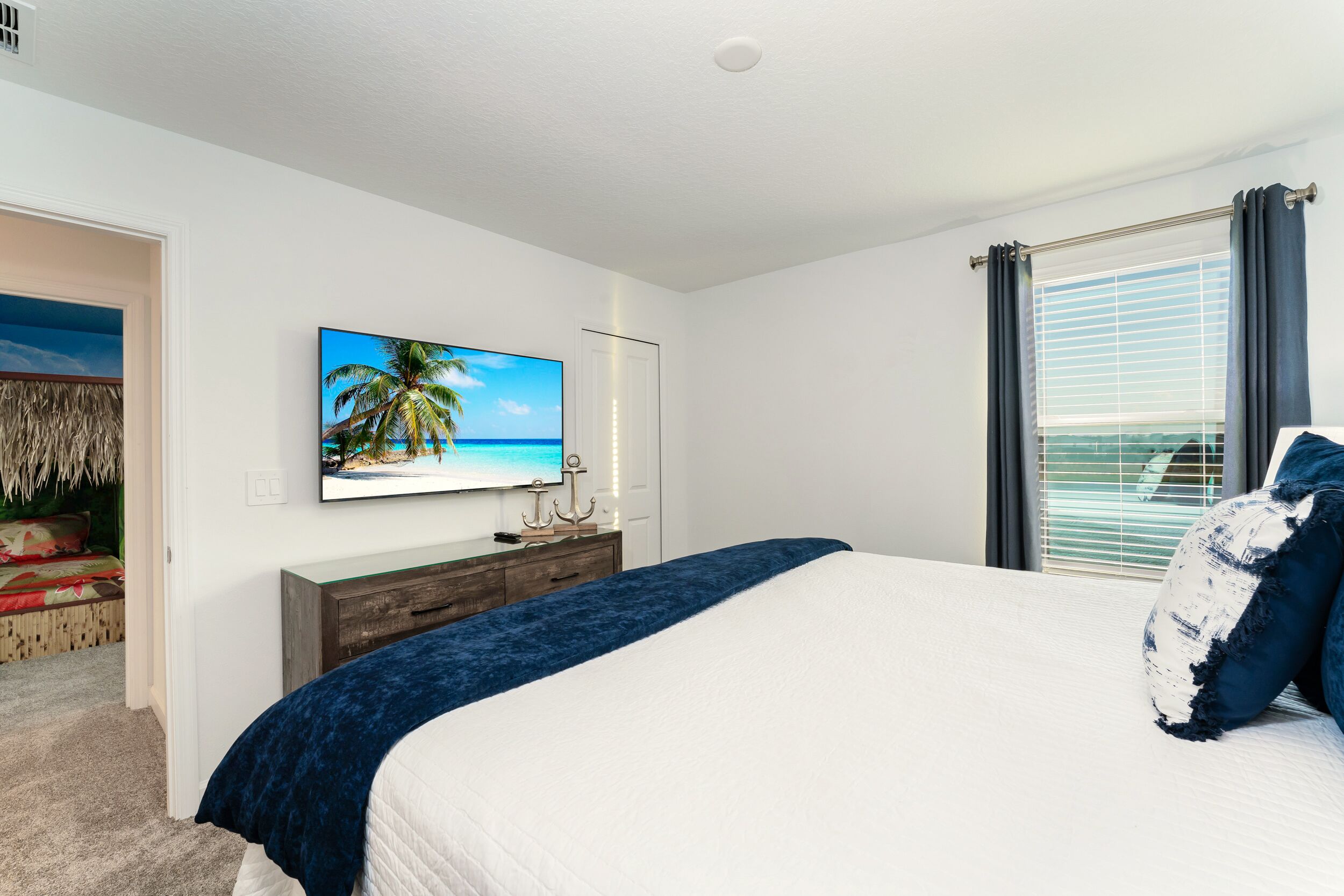 A bedroom with a bed, a television on the wall, a window with curtains, and a door leading to another room.