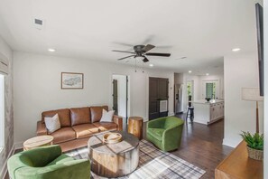 A spacious living area with a brown leather sofa, green armchairs, a round coffee table, and a ceiling fan. A kitchen is visible in the background.