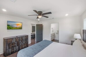 A bedroom with a bed, a ceiling fan, a dresser, a desk, and a bathroom visible through an open door.