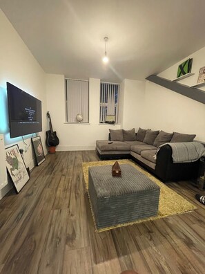 A living room with a sectional sofa, a flat-screen TV, a guitar, and a small table with a globe. - Modern 1 bed Flat- 20mins From Central London (Romford)