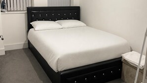 Ein Einzelbett mit schwarzem Kopfteil und weißer Bettwäsche in einem Zimmer mit grauem Teppich, einem weißen Heizkörper und einem Fenster mit Jalousien.