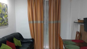 Un salon avec un canapé en cuir noir, des coussins verts et rouges, une table à manger avec des chaises en bois et des rideaux à rayures.