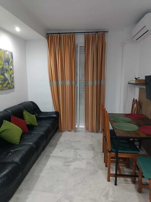 Ein Wohnzimmer mit einem schwarzen Ledersofa, grünen und roten Kissen, einem Esstisch mit Holzsesseln und gestreiften Vorhängen.