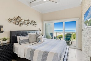 Une chambre à coucher comprenant un lit, une table de chevet, une lampe fixée au mur, un ventilateur de plafond et une vue sur la plage, accessible par une porte-fenêtre coulissante.