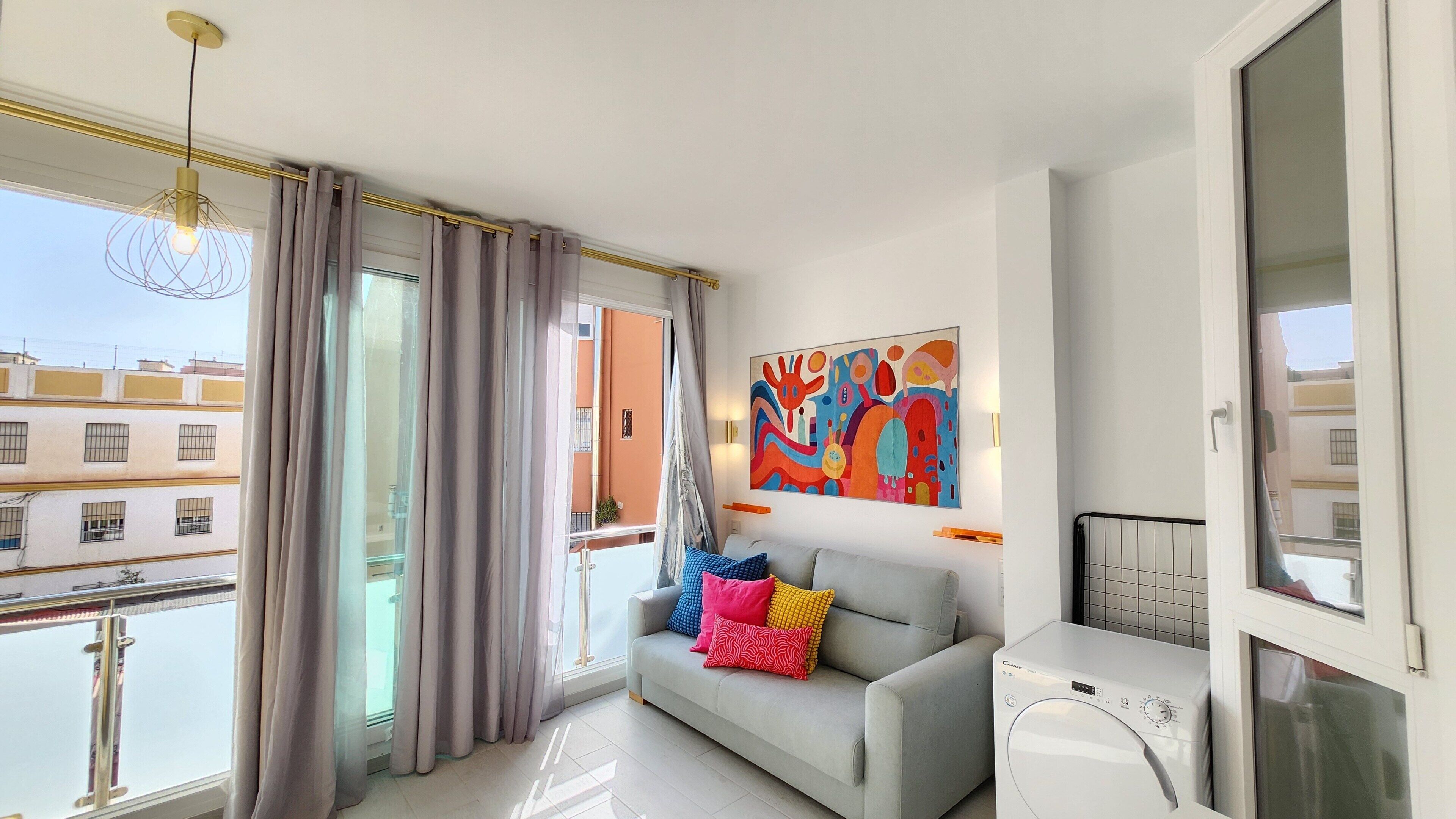 Een moderne woonkamer met een grijze bank, kleurrijke kussens, een groot abstract schilderij, een wasmachine en een balkon met gordijnen.