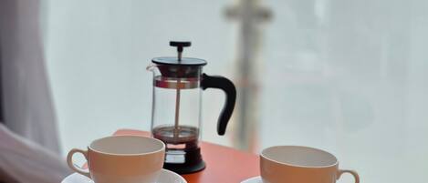 A table with two white cups and a French press on a wooden surface, with a window in the background.