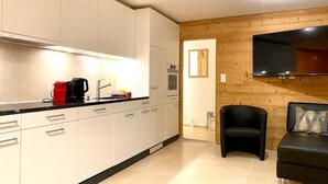 A modern kitchen with white cabinets, a black countertop, and a black sofa in a living area with wooden walls and a flat-screen TV.