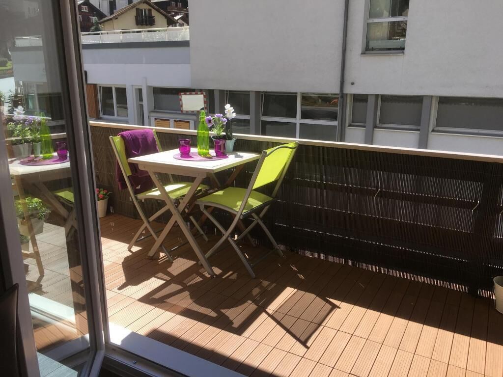 Un balcón con una mesa, sillas, una planta en maceta y una ventana con vista a un edificio.