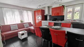 A modern kitchen with red cabinets, a dining area with red chairs, and a sofa with grey cushions.