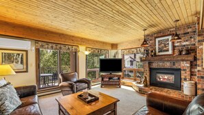 A cozy living room with a fireplace, leather furniture, a television, and a large window overlooking greenery. - Slope-Side Retreat at the Steamboat Resort Base Area (Steamboat Springs)