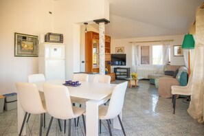 A dining area with a white table and chairs, a kitchen with a refrigerator and oven, a living room with a sofa and a TV, and a window with curtains. - Solemar Sicilia -Residence Vara (Piana Calzata)