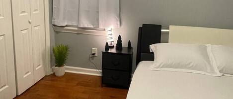 A bedroom with a bed, a nightstand, a potted plant, a window with curtains, and a door.