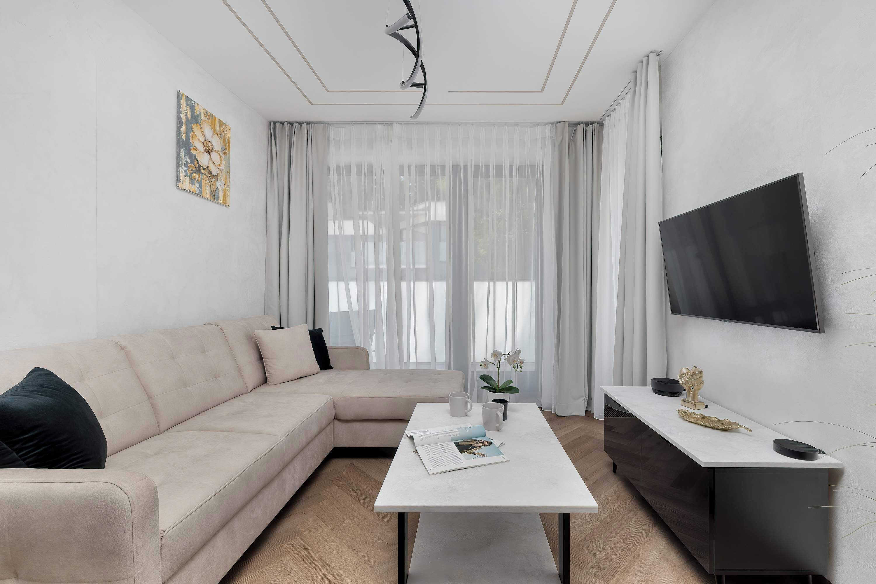 A bright and modern living room features a comfortable beige corner sofa and a sleek wall-mounted television. A minimalist white coffee table sits in the center on a light wood-patterned floor.