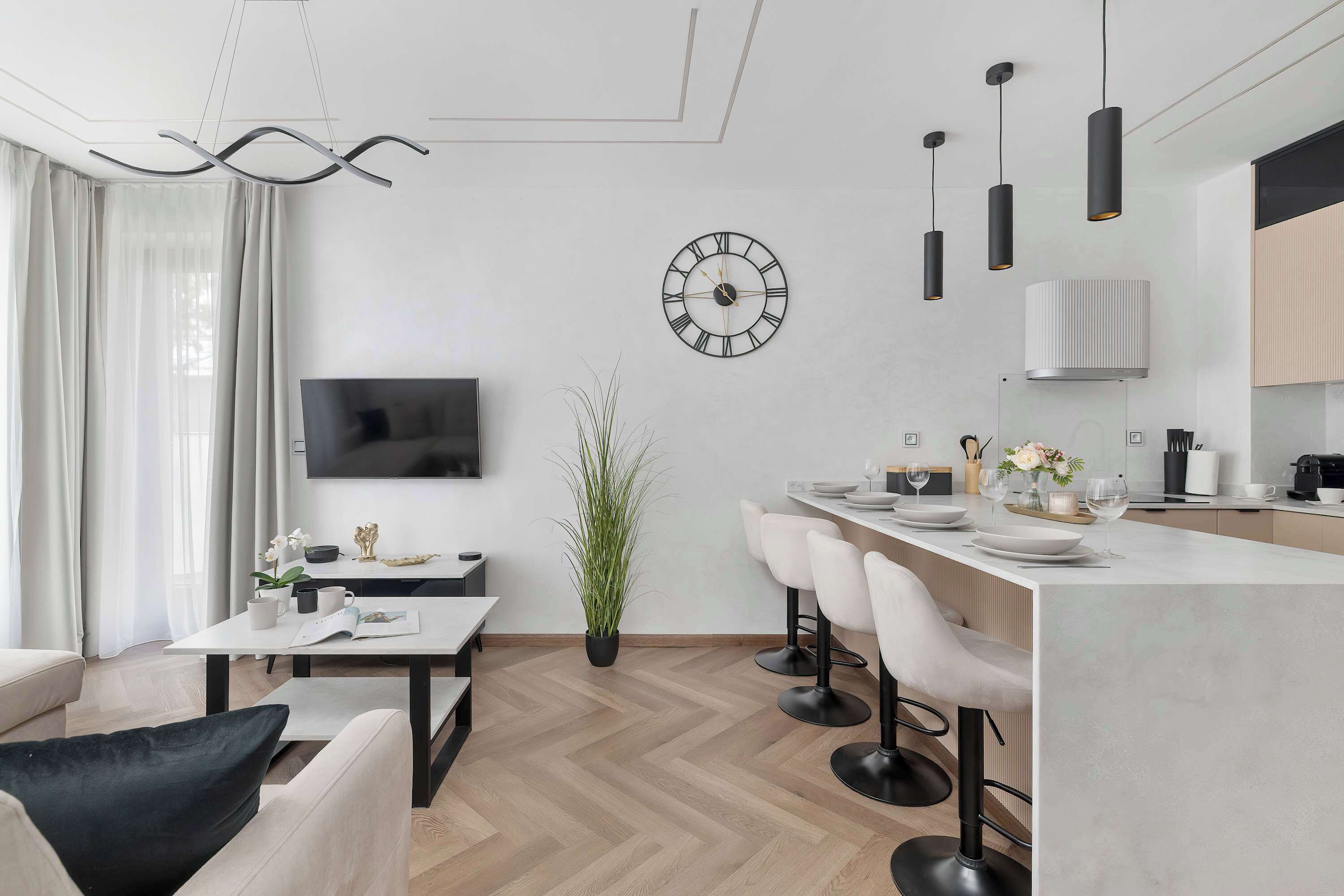 A modern kitchen with a central island, bar stools, a flat-screen TV, and a wall-mounted clock.