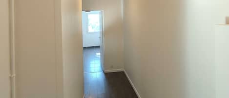 A narrow hallway with white walls and wooden flooring, leading to a door at the end.