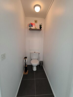 Une petite salle de bain comprenant des toilettes, une étagère avec des articles de toilette et une petite poubelle.