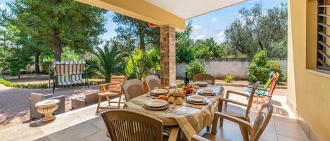 Un área de comedor al aire libre con una mesa lista para cuatro personas, rodeada de sillas, un pilar de piedra y vistas a un jardín con árboles y una valla.
